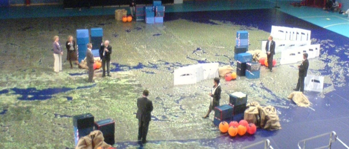 Politicians and officials standing on a hall-sized map of the Mälardalen region, moving boxes, sacks and markers across it as game pieces
